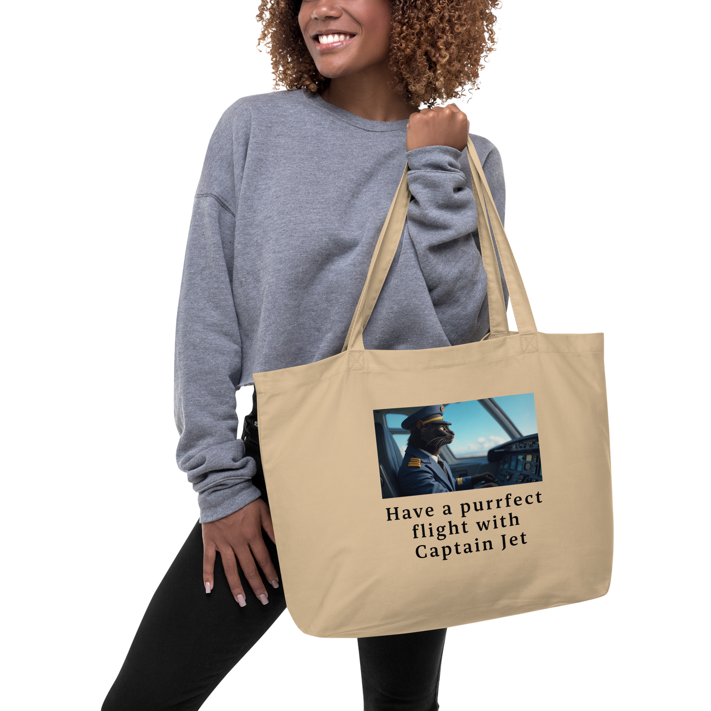 Woman holding a beige large tote bag. Organic cotton. With a print of a black cat in a plane. Wearing a pilot's uniform and text.