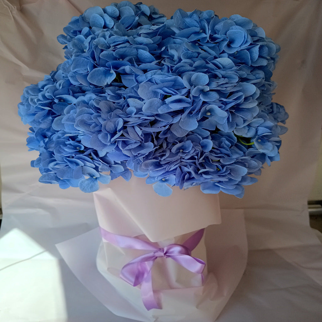 A large bouquet of pretty blue artificial hydrangeas. In a glass vase and wrapped with paper and a ribbon.