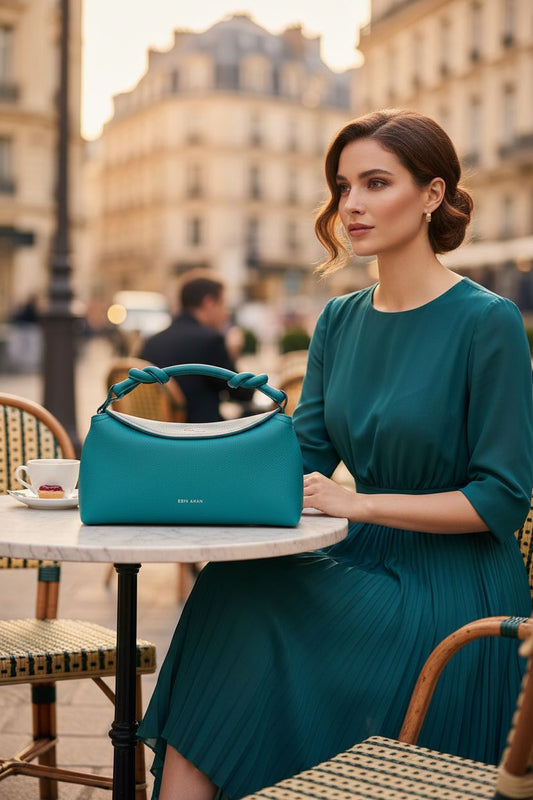 Woman sitting at outdoor café with Midi Sloane teal leather crossbody bag on table. Made by artisans.