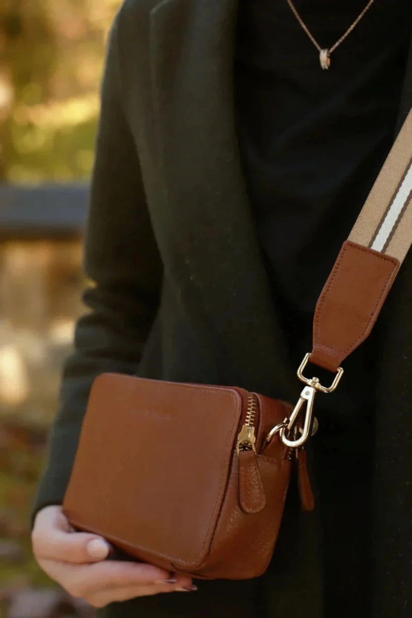 Woman with a Bayraw brown leather crossbody bag with gold zips and clasps. Brown stripe strap. 
