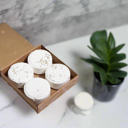Four white shower steamers in a cardboard box on a marble surface. A plant next to the box.