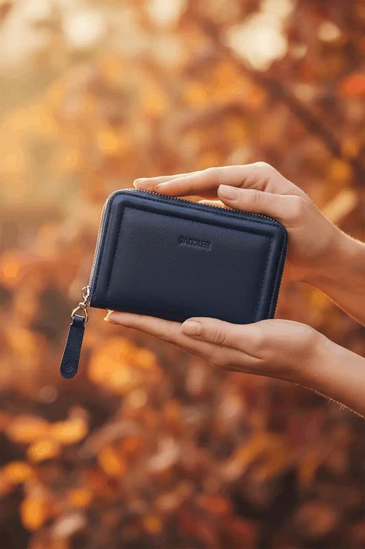 A woman holding a navy blue Saddler Annie real leather zip-around wallet in two hands. Blurred background of autumn leaves.